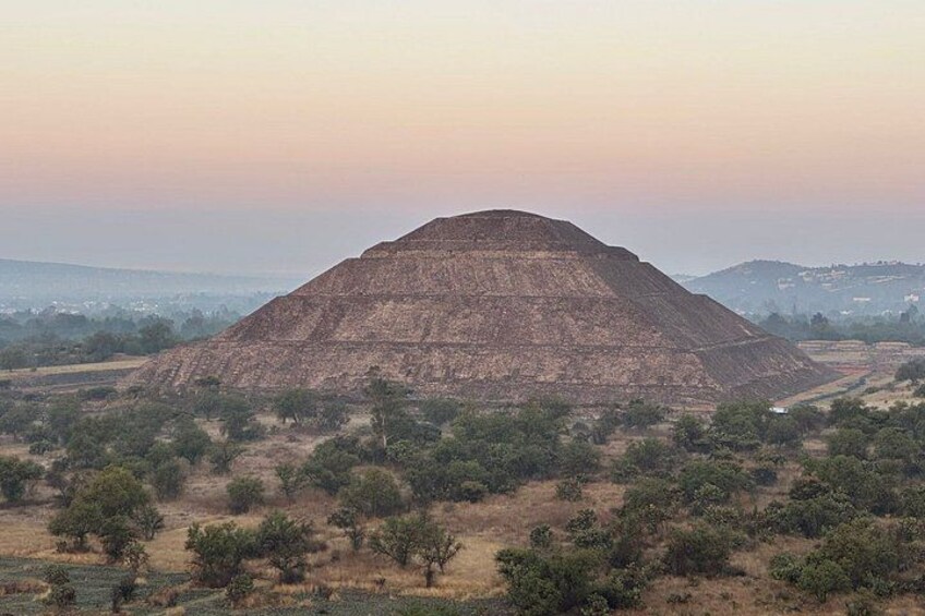 A large, flat-topped pyramid-like structure in a rural landscape with sparse trees and distant hills. 3