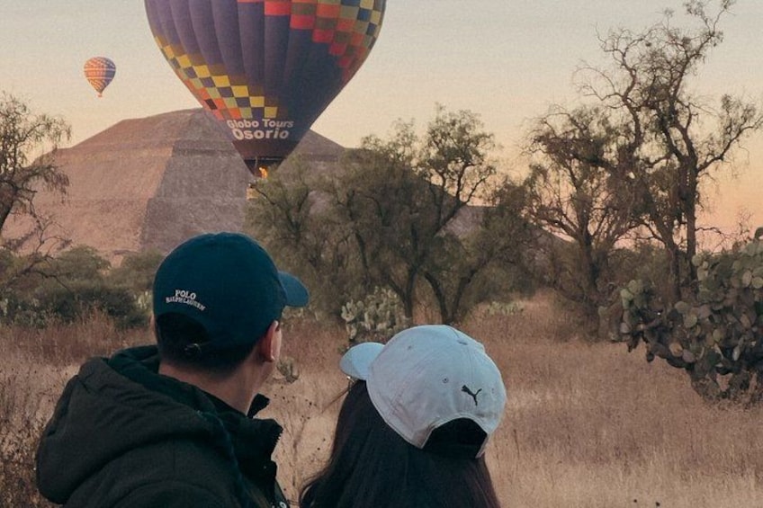Two people observing a hot air balloon in a desert landscape with sparse vegetation and a mountain in the background. 1
