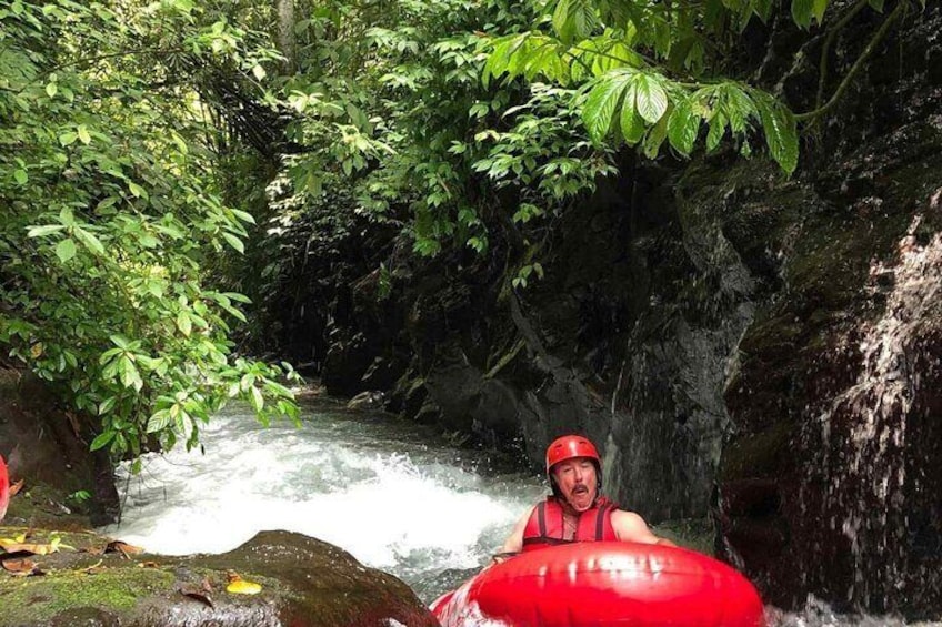 A person in a red helmet and life jacket on a red raft navigating a river surrounded by dense foliage. 1