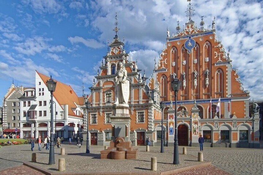 En historisk bygning med detaljeret arkitektur, et brostensbelagt torv og en statue i midten. 2