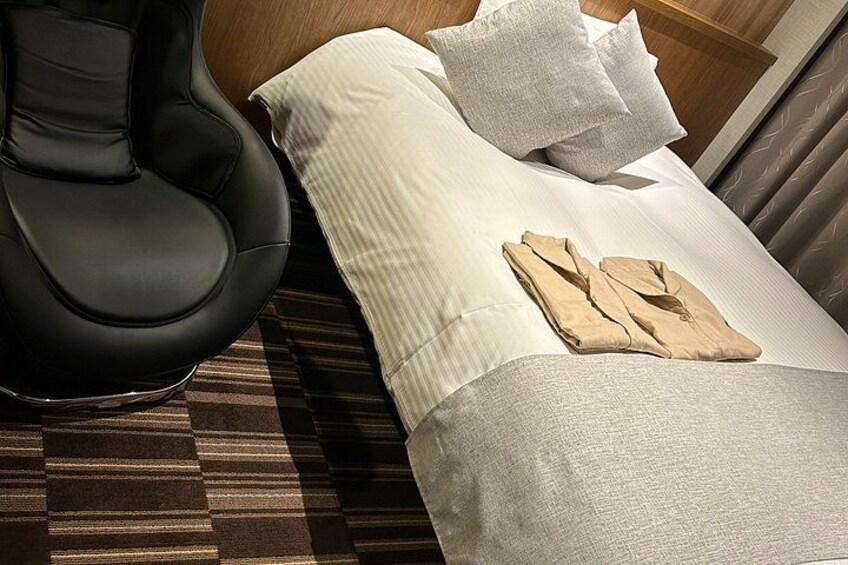 A bed with white bedding, a black massage chair, and a folded beige jacket on the bed. 5