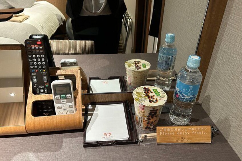 A hotel room with a wooden side table holding a remote control, a smartphone, a notepad, a pen, a cup of bubble tea, and two bottles of water. 3