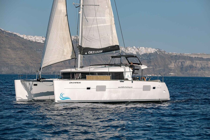 A white sailboat with 'Okeanos' written on its sails and hull, sailing on a body of water with a mountainous coastline in the background. 5