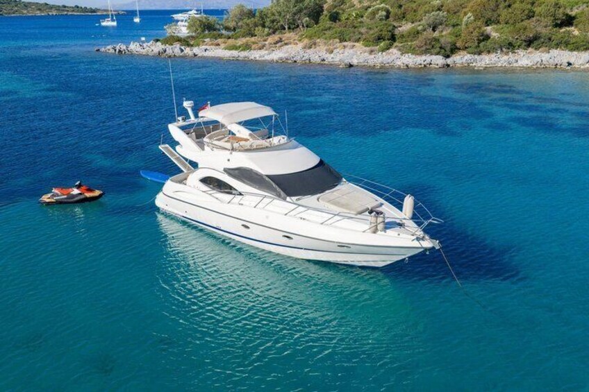A white yacht anchored in clear blue waters with a rocky shoreline and sparse vegetation in the background. 1