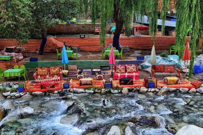 A riverside seating area with colorful cushions and tables, surrounded by trees and a flowing stream. 2