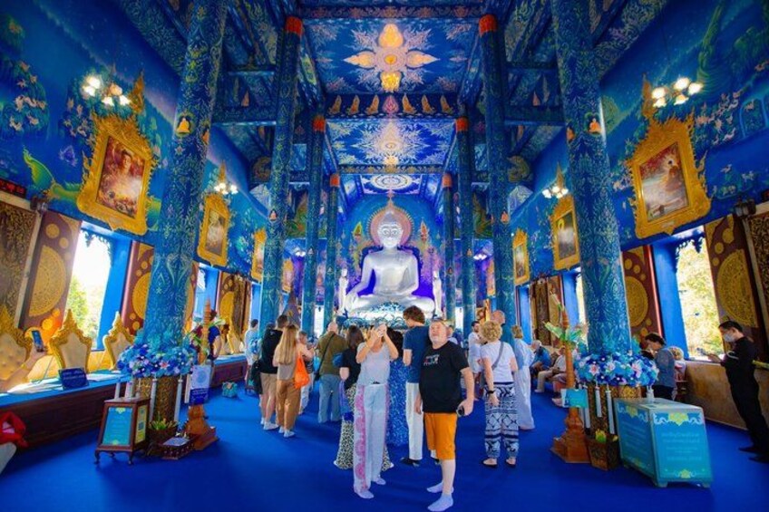A temple interior with a central Buddha statue, ornate blue and gold decorations, and visitors. 2