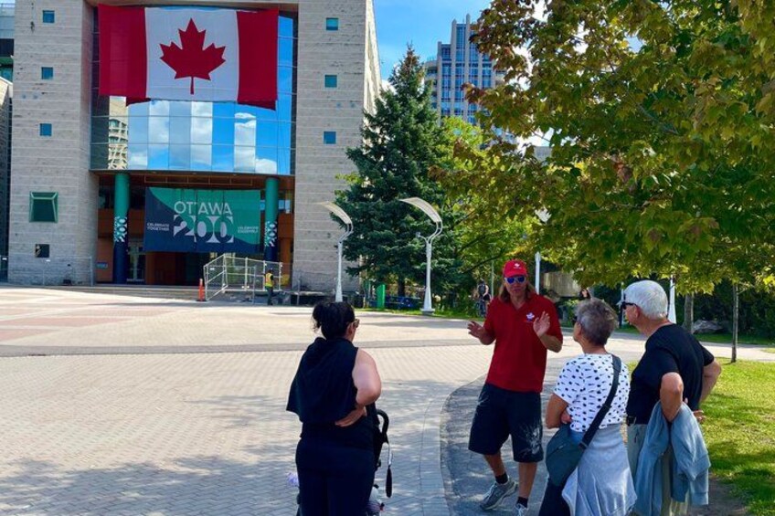 A group of people standing in a park with a large building featuring a Canadian flag and a banner for the 200th anniversary of Ottawa. 4