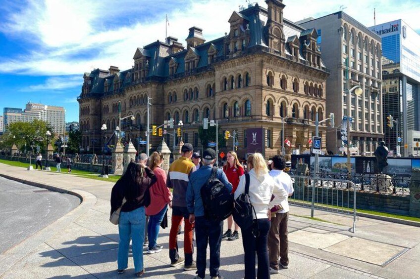 A group of people standing on a sidewalk in front of a historic building with arched windows and a clock tower. 2