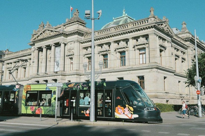A tram at a bus stop in front of a large, ornate building. 2