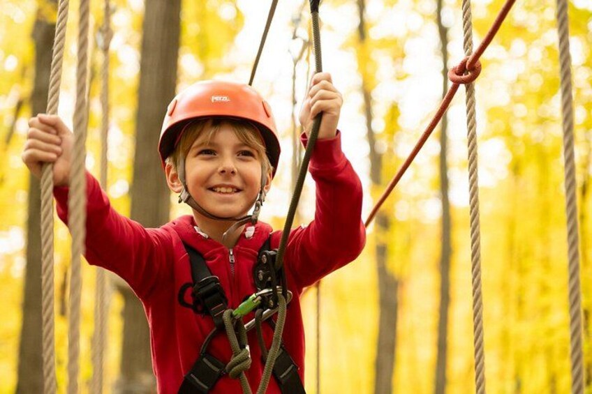 A child in a red jacket and helmet on a rope course, surrounded by trees. 2