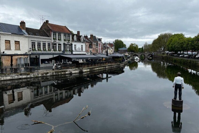 A calm river reflecting buildings, a person standing on a dock, and a bridge in the distance. 5
