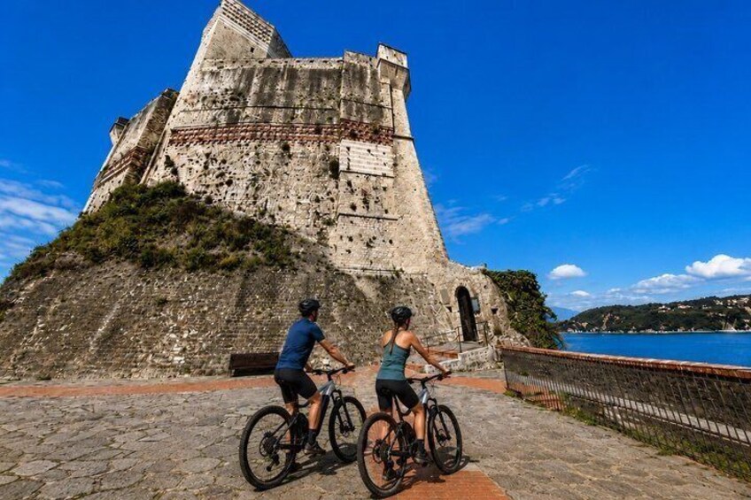 Two cyclists on a cobblestone path with a historic stone wall and a body of water in the background. 2