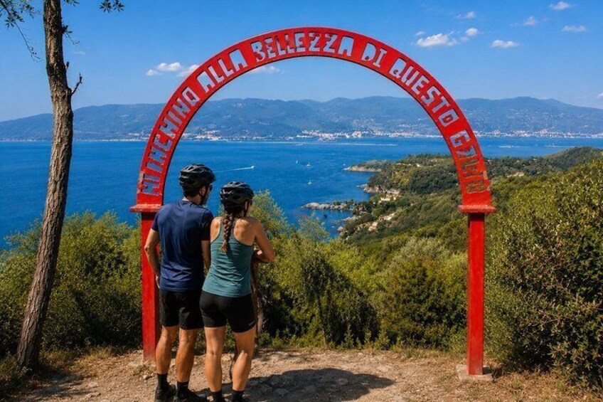 A couple standing under a red archway with a scenic view of the sea and hills in the background. 4