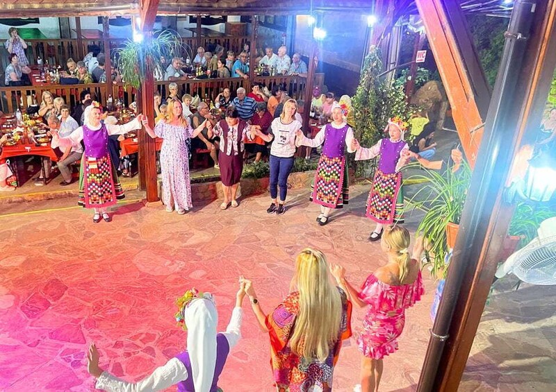 A group of people in traditional attire performing a dance on a stage with an audience seated in the background. 1