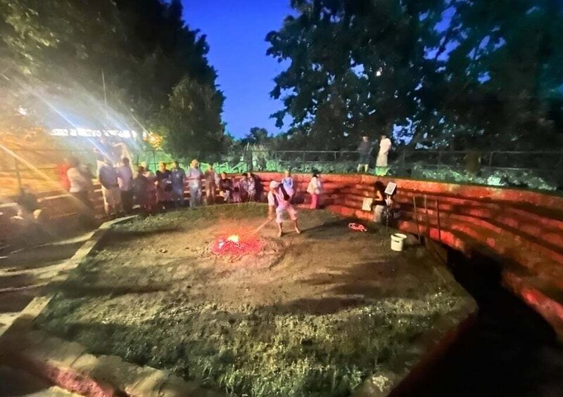 A nighttime outdoor event with a crowd gathered around a central fire, surrounded by a brick wall and trees. 3