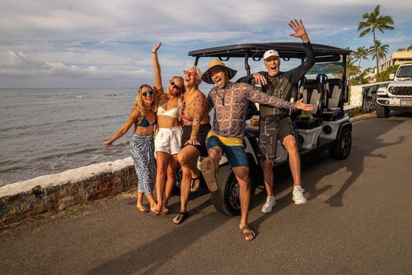 A group of people posing by a coastal road with a golf cart and a vehicle in the background. 2