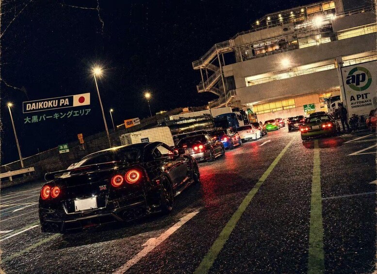 A nighttime urban scene with a black sports car, a building with 'APIT AUTOBACS SHINONOME' signage, and a 'DAIKOKU PA' sign. 1