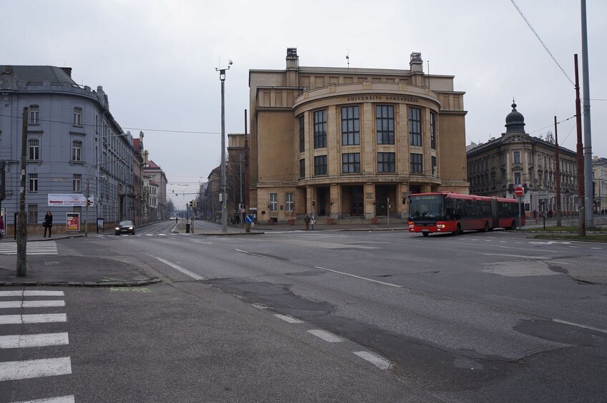 Una calle con un autobús rojo, un paso de peatones y un edificio con la inscripción 'UNIVERSZITA KOMENSKIEGO'. 2