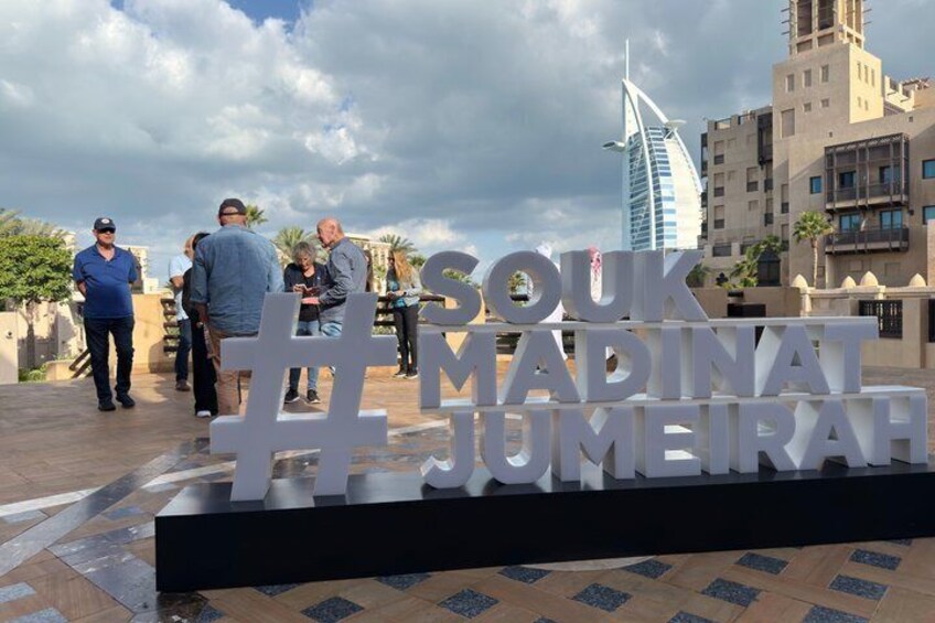 A sign with the text 'SOUK MADINAT JUMEIRAH' in a public square with people around. 4