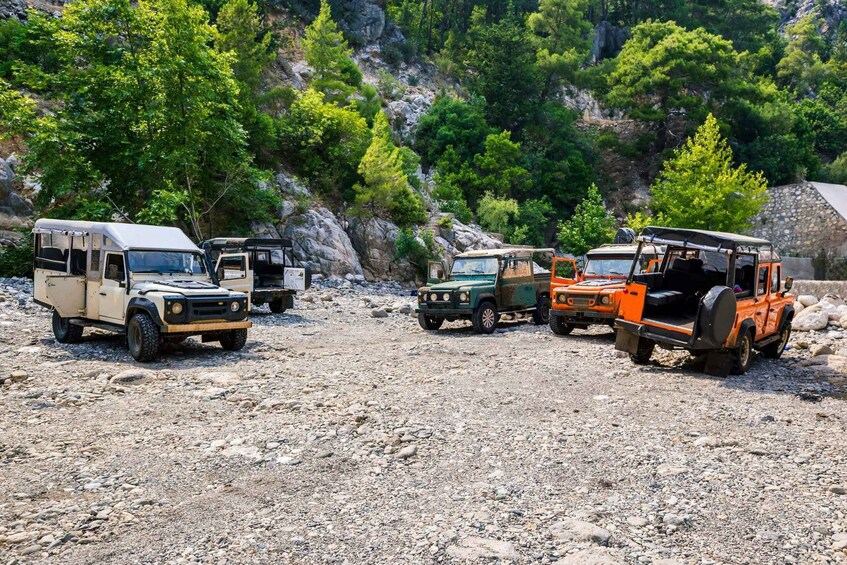 A group of off-road vehicles parked on a rocky terrain with a backdrop of dense green trees and a stone wall. 3