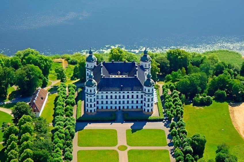 An aerial view of a large, historic building with multiple towers, surrounded by lush greenery and a body of water. 1