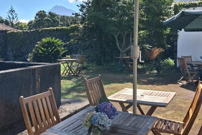 A wooden table with chairs and a flower arrangement in a garden with a stone wall and trees. 1