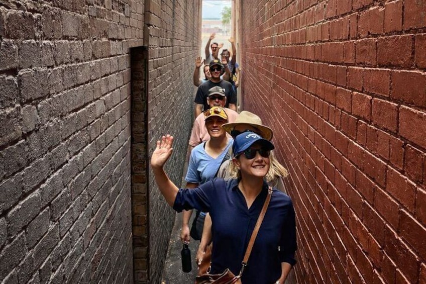 A narrow alley with brick walls, a person in the foreground waving, and others in the background. 1
