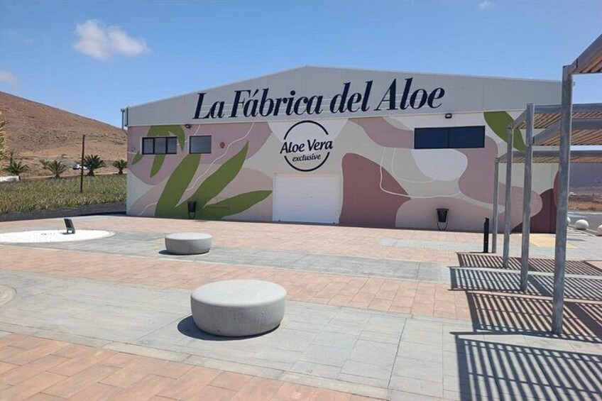 A building with a sign reading 'La Fábrica del Aloe' and 'Aloe Vera Exclusive.' There are outdoor seating areas with round tables and benches. 3