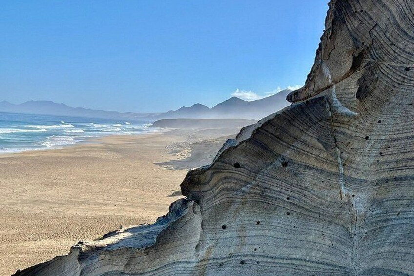 A coastal landscape with a prominent rock formation, a sandy beach, and distant mountains under a clear sky. 1
