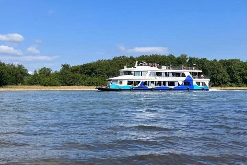 A large, multi-deck riverboat with a blue and white color scheme, floating on calm water with a forested shoreline in the background. 1