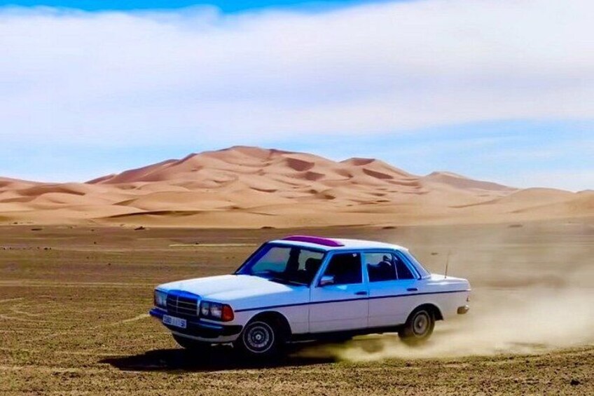 A white car driving on a sandy terrain with hills in the background. 1
