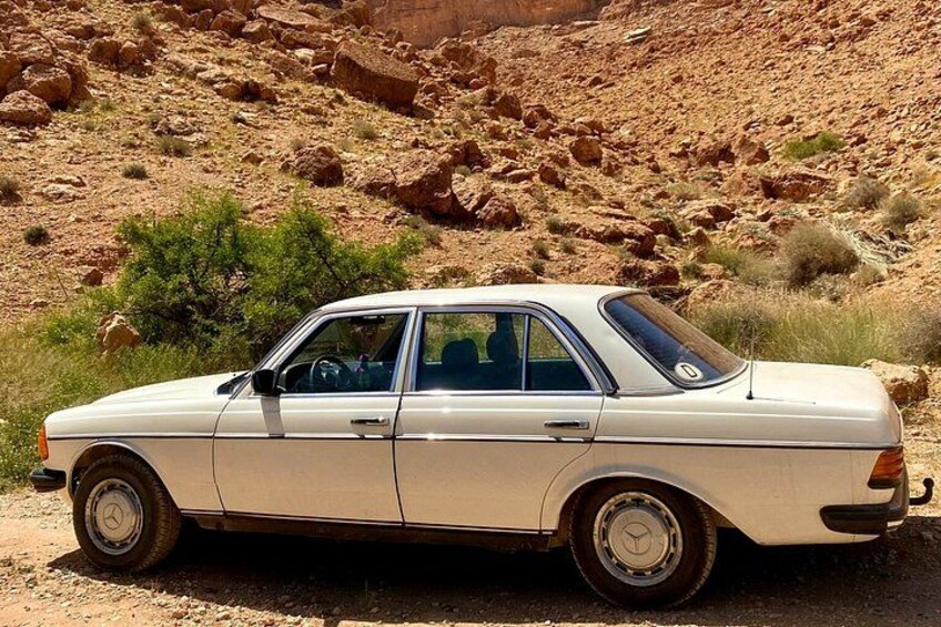A vintage beige car parked on a dirt road with rocky terrain and sparse vegetation in the background. 4