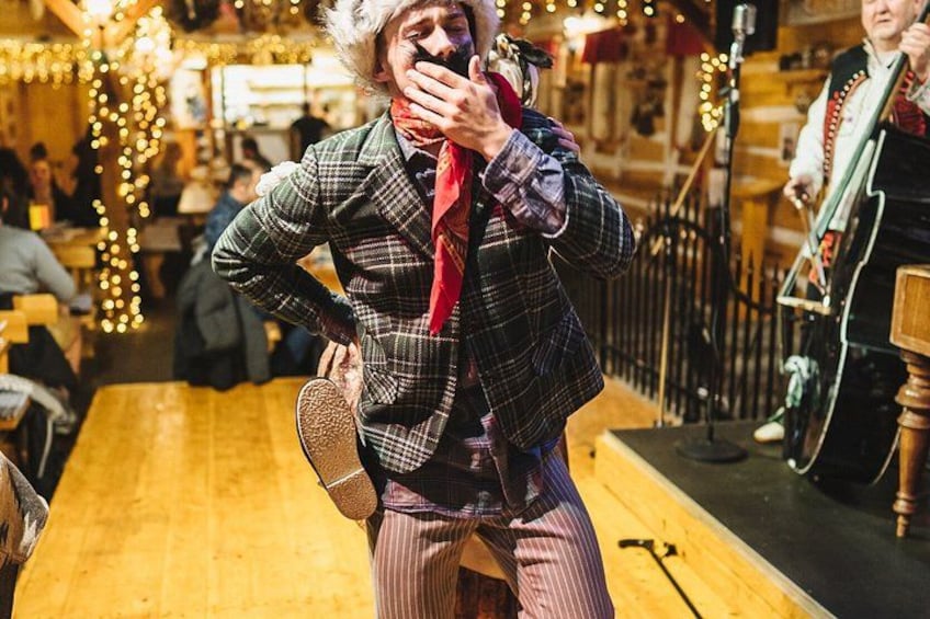 A person in a plaid jacket and striped pants, holding a microphone, in a warmly lit indoor setting with wooden floors and string lights. 5