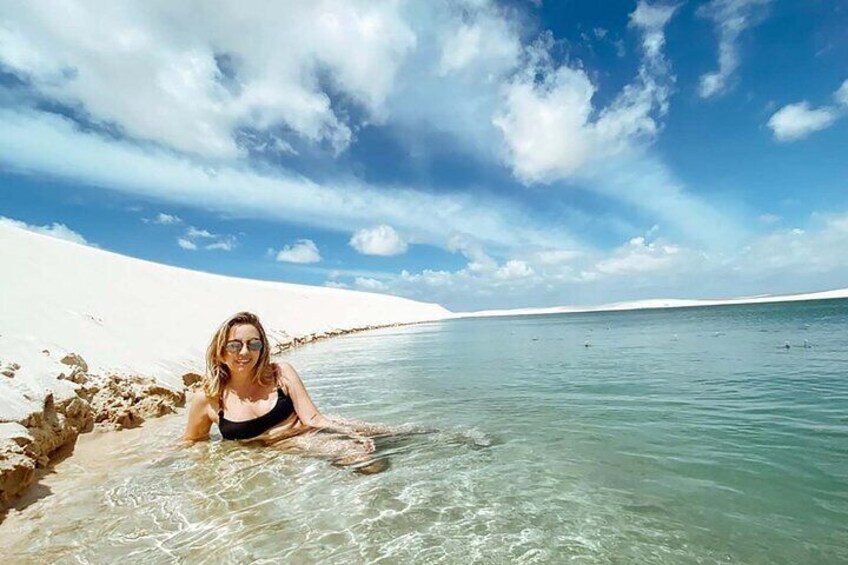 A person in a black swimsuit lying on a sandy beach with clear turquoise water and white sand dunes in the background. 3