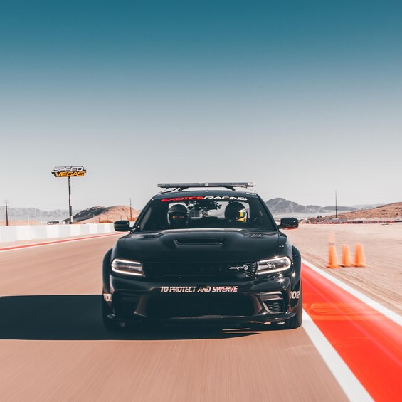 A black racing car with 'ExoticsRacing' and 'To Protect and Serve' decals on a desert road with orange traffic cones. 5