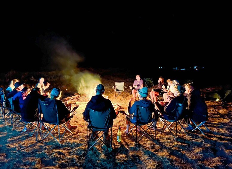 A group of people sitting in chairs around a campfire at night. 3