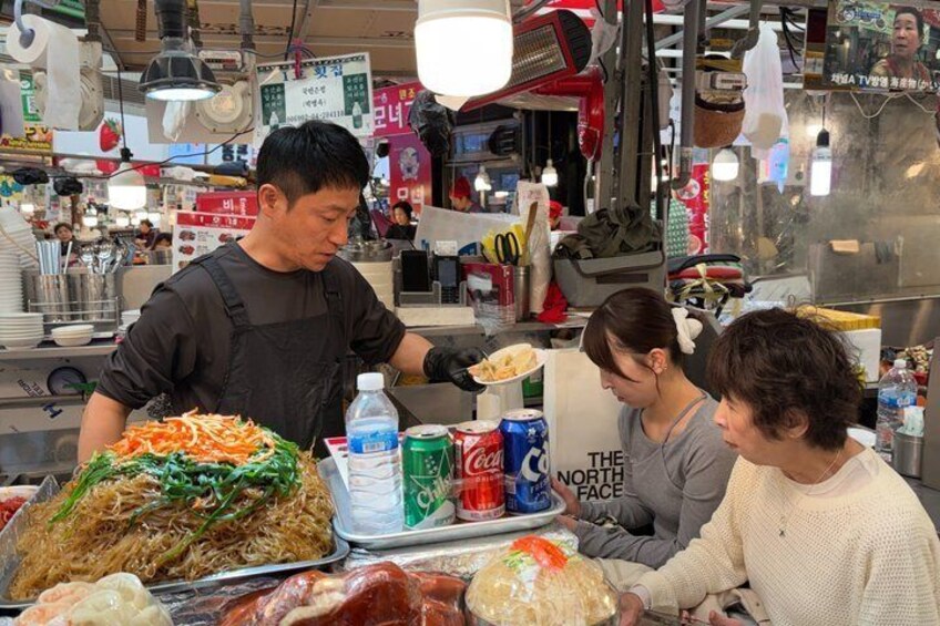 A street food vendor preparing noodles with vegetables, a customer, and a woman in the background. 4