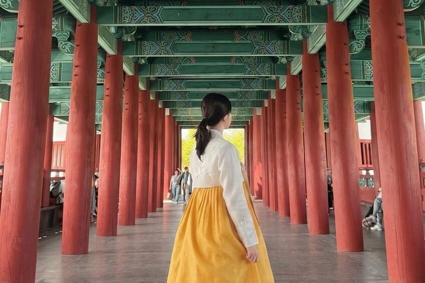 A traditional Korean hanbok, a woman in a yellow skirt and white blouse, walking through a corridor with red wooden pillars and green ceiling decorations. 2