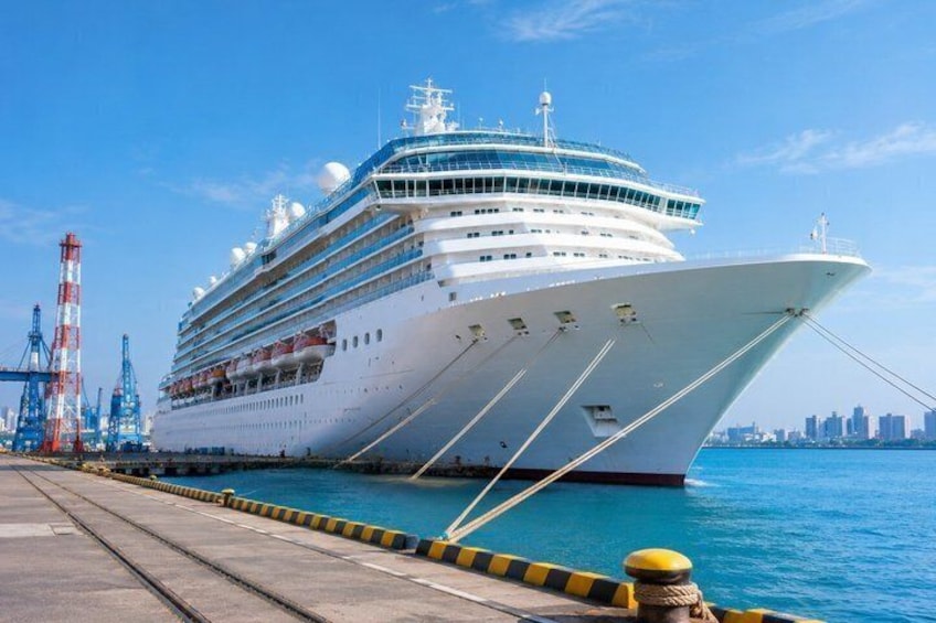 A large cruise ship docked at a port with cranes and a city skyline in the background. 1