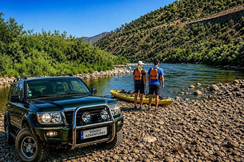 A black SUV parked by a river with two people in life jackets and a yellow kayak. 1