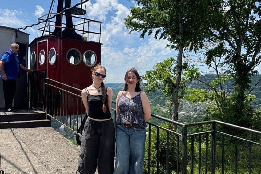 Two people standing on a platform with a red structure featuring circular windows, overlooking a landscape with trees and hills. 2
