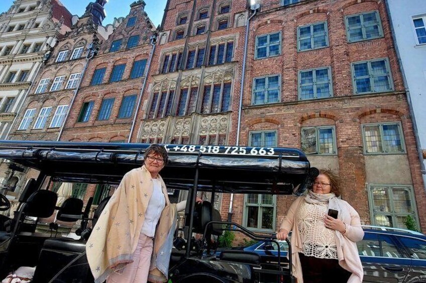 Two people standing in front of a historic building with a black bus and a car parked nearby. 4