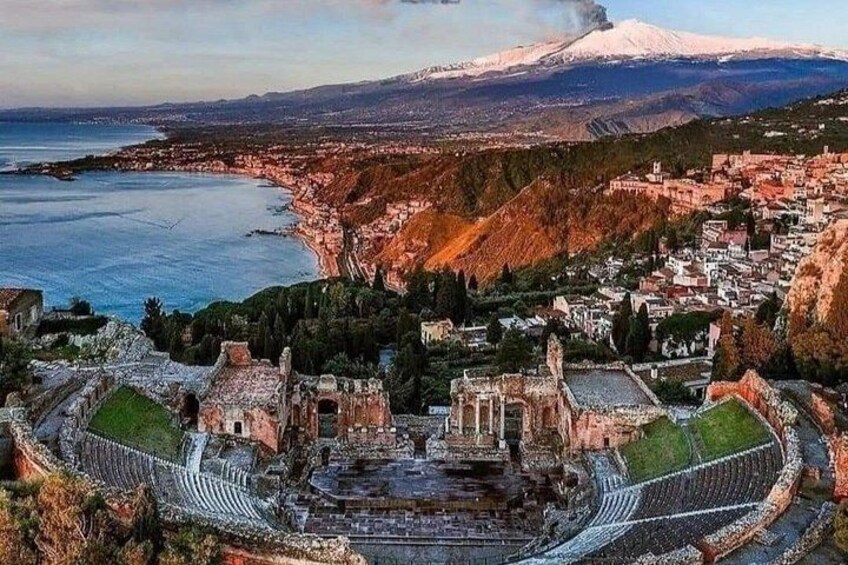 An ancient amphitheater with ruins, a coastal town, and a snow-capped mountain in the background. 1