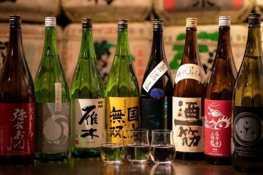 A variety of Japanese sake bottles with labels, some with glasses in front. 1