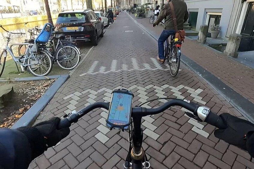 A bicycle with a GPS device, a paved path, parked bicycles, and a person riding a bicycle in the background. 1