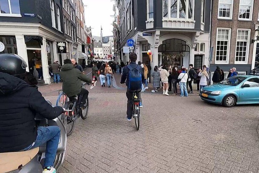 A cobblestone street with cyclists and pedestrians, historic buildings, and a blue car. 2