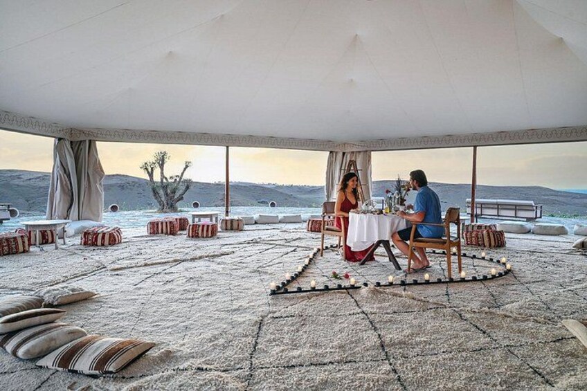 A couple dining under a tent on a rocky terrain with cushions and a table set for two. 1
