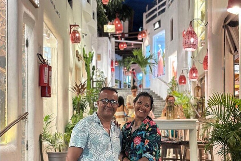 A couple standing on a narrow street with buildings on either side, red lanterns, and potted plants. 1