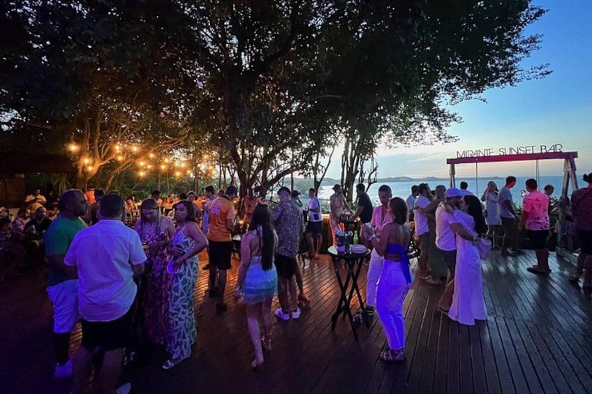 A crowded outdoor bar with people socializing, a wooden deck, and a view of the ocean. 3