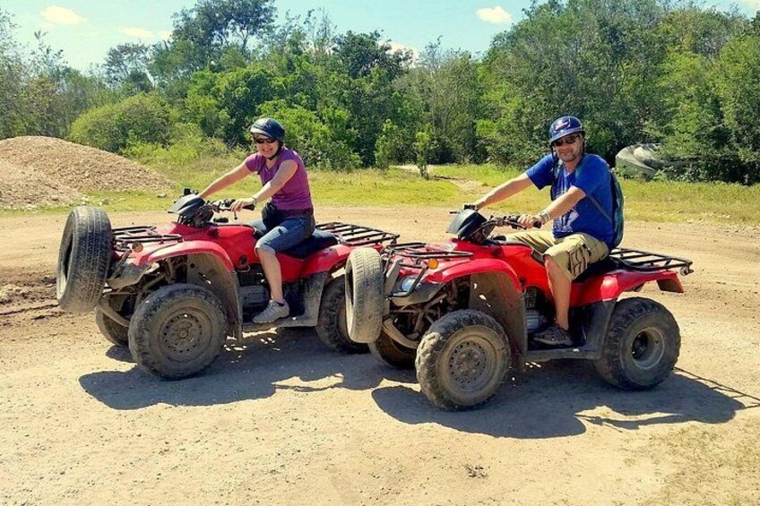 Two people on red all-terrain vehicles on a dirt path with trees in the background. 1